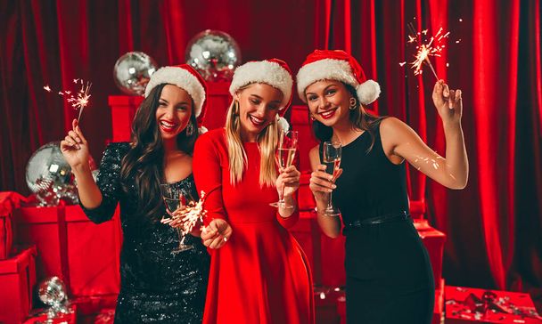 Happy New Year to you! Three beautiful sexy women in Santa hats with glasses of champagne and sparkles. New Year's party. Christmas Eve. Against the background of red gift boxes and disco balls. - Fotó, kép