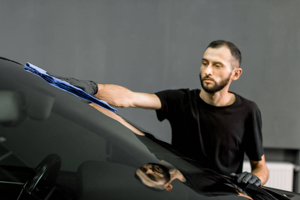 Car wash and cleaning at professional auto service station. Shot of handsome bearded young male worker in black t-shirt and gloves, cleaning car windscreen with blue microfiber cloth - Foto, afbeelding