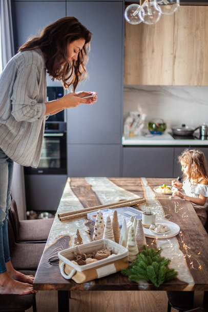 Young woman standing on chair and making top view photos of Christmas composition in the kitchen while her daughter eating breakfast. Concept of using technology for taking photos for social media. - Φωτογραφία, εικόνα