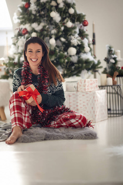 Egy fiatal vidám lány otthon pózol egy fotón egy karácsonyi ajándékkal egy gyönyörű ünnepi reggelen. Karácsony, kapcsolat, szerelem, együtt - Fotó, kép