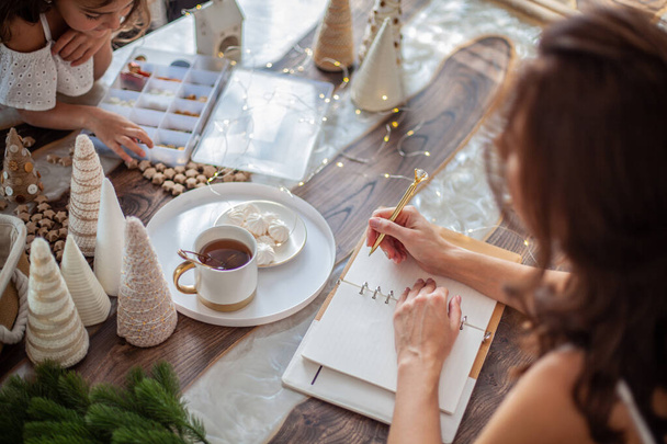 Young woman drinking tea and writing plans or goals for New Year 2021 while her daughter crafting Christmas trees from paper cone, yarns and buttons with placed stars and fairy lights on wooden table. - Foto, Imagen
