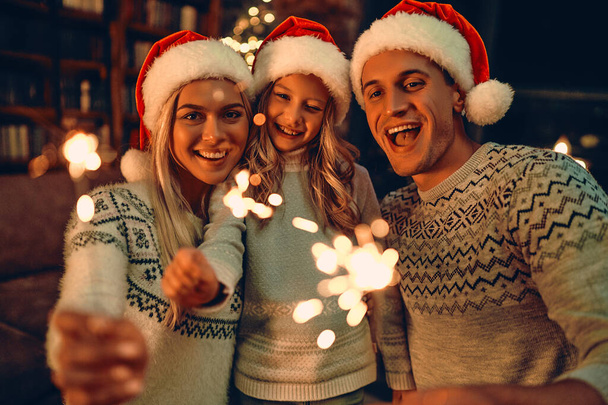 Feliz Natal e Feliz Ano Novo! Família feliz celebrando férias de inverno em casa. Pais e sua filha esperando o Natal em chapéus de Papai Noel com faíscas nas mãos. - Foto, Imagem