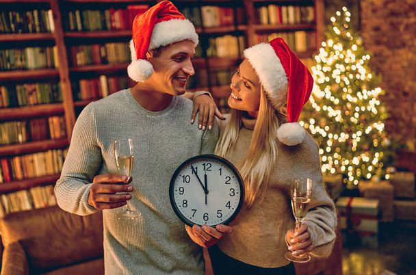 Καλά Χριστούγεννα και Καλή Χρονιά! Νεαρό ζευγάρι με ρολόι στα χέρια χαμογελώντας και κοιτάζοντας την κάμερα. Πέντε λεπτά μέχρι την Πρωτοχρονιά.. - Φωτογραφία, εικόνα