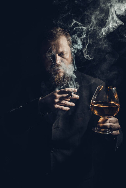 Solid confident bearded man in suit with glass of whisky and cigar with fume - Foto, Bild