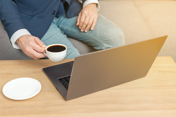 Unrecognized man, businessman or freelancer sitting on sofa, drinks coffee and working at laptop, work from home. - Fotoğraf, Görsel