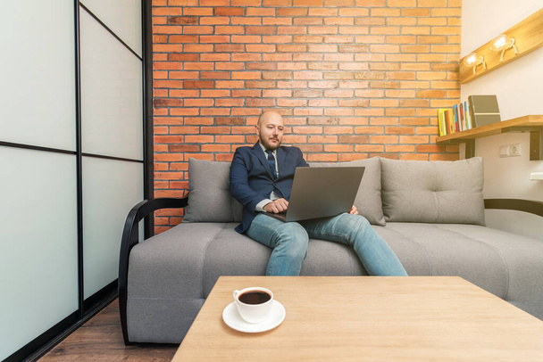 Bearded bald man, businessman or freelancer sitting on sofa and working on laptop from home, modern interior loft design. - Photo, Image