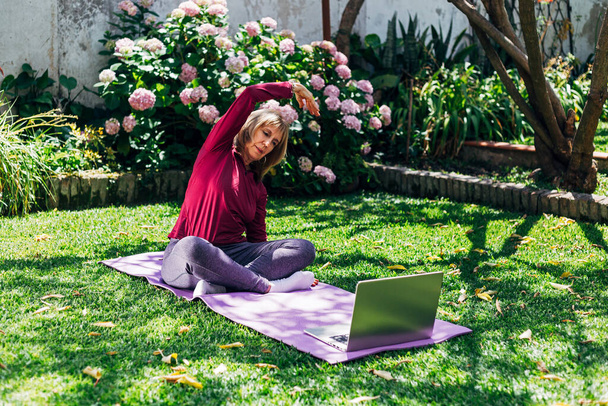 Онлайн тренировки yoga спорта, старшая женщина в саду ее дома делать упражнения на циновке пилатеса напротив ноутбука на дому. Тренер на видеоконференции. Понятие здорового образа жизни. - Фото, изображение