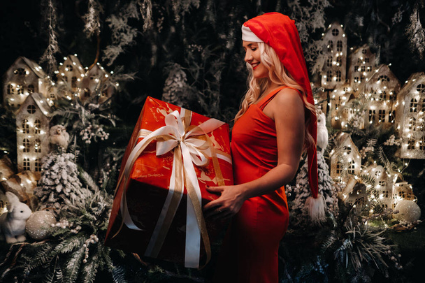 a girl in a Santa hat with a big Christmas gift in her hands on a fabulous Christmas background.A smiling woman in a red dress on the background of Christmas trees and small houses. - Fotoğraf, Görsel