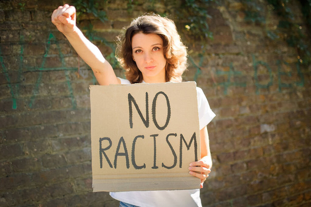 Upset young protesting woman in white shirt holds protest sign broadsheet placard with slogan No racism for public demonstration on stone wall background - Photo, Image