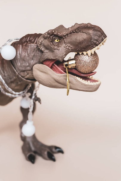 Tyrannosaure jouet du Nouvel An avec une guirlande sur le cou et une décoration d'arbre de Noël dans les dents - Photo, image