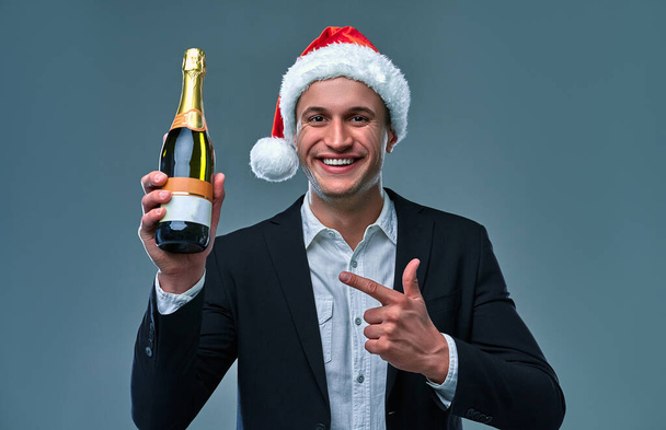 Successful man in a jacket and Christmas hat points a bottle of champagne celebrates New Year. Studio photo on a gray background . - Foto, Imagen