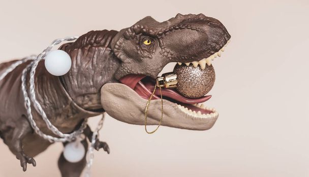 Новорічна іграшка тиранозавр з гірляндою на шиї і прикрасою ялинки в зубах
 - Фото, зображення
