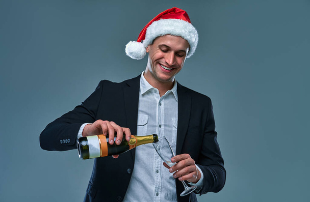 Succesvolle man in een jasje en kerstmuts giet champagne in een glas viert Nieuwjaar. Studio foto op een grijze achtergrond . - Foto, afbeelding