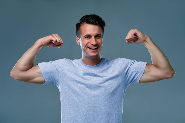 Portret van sportieve, aantrekkelijke, krachtige, viriele, harde, sexy man in t-shirt met opgeheven armen, met reliëfspieren op zijn handen, geïsoleerd op grijze achtergrond - Foto, afbeelding