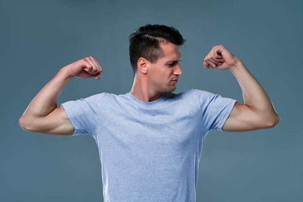 Porträt eines sportlichen, attraktiven, kraftvollen, virilen, rauen, sexy Typen im T-Shirt mit erhobenen Armen, mit Entlastungsmuskeln an den Händen, isoliert auf grauem Hintergrund - Foto, Bild