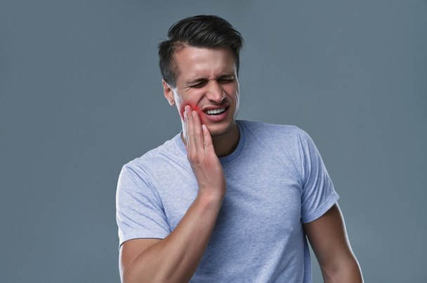 Концепция зубной боли. Внутренний снимок молодого мужчины, чувствующего боль, держащего щеку за руку, страдающего от сильной зубной боли, смотрящего в камеру с болезненным выражением лица - Фото, изображение