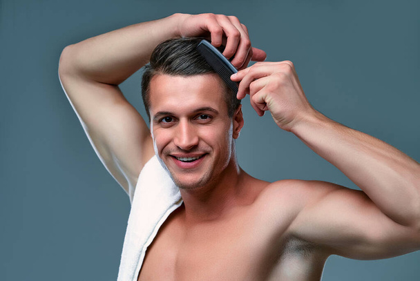 Handsome young man isolated. Portrait of shirtless muscular man with a towel on his shoulder is standing on gray background and combing out his hair. - Fotoğraf, Görsel