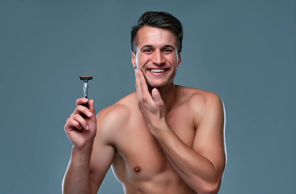 Handsome young man isolated. Portrait of shirtless muscular man is standing on grey background with razor in hand and smiling. Men care concept. - Photo, Image