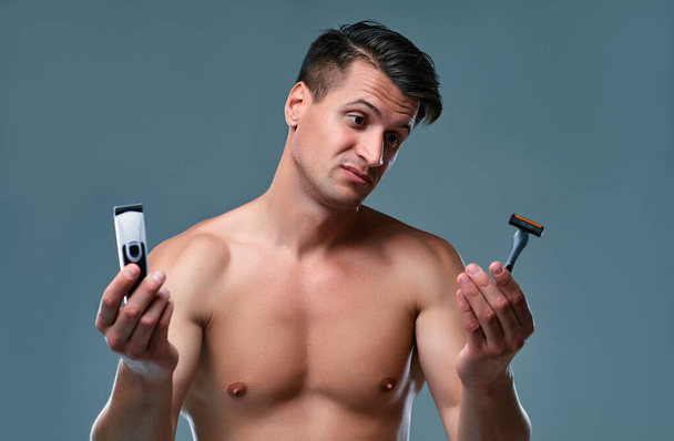 Handsome young man isolated. Portrait of shirtless muscular man is standing on grey background with trimmer in one hand and razor in other. Men care concept. - Φωτογραφία, εικόνα