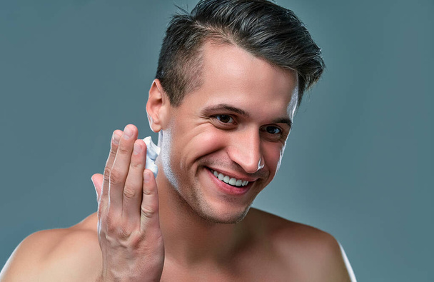 Atrakcyjny mężczyzna na szarym tle używa pianki do golenia na twarzy. Opieka nad mężczyznami. - Zdjęcie, obraz