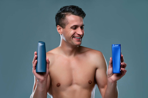 Handsome young man isolated. Portrait of shirtless muscular man is standing on grey background with shampoo in hands. Men care concept. - Foto, imagen