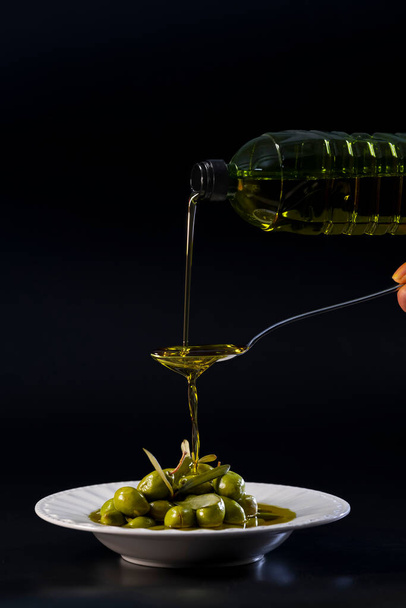 Оливковое масло выливается из пластиковой бутылки в ложку и падает в тарелку с оливками - Фото, изображение