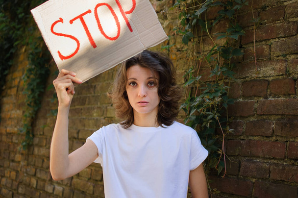Молода жінка, що протестує у білій сорочці та джинсах, тримає знак протесту, плакат з гаслом Стоп для публічної демонстрації на фоні стіни
. - Фото, зображення