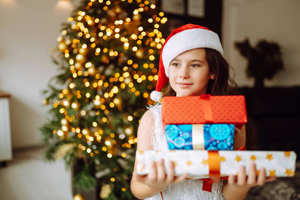 Happy female child in Santa red hat holding Christmas gifts. Smiling little girl posing with many presents in Christmas interior design. Winter holiday, New Year. - Foto, imagen