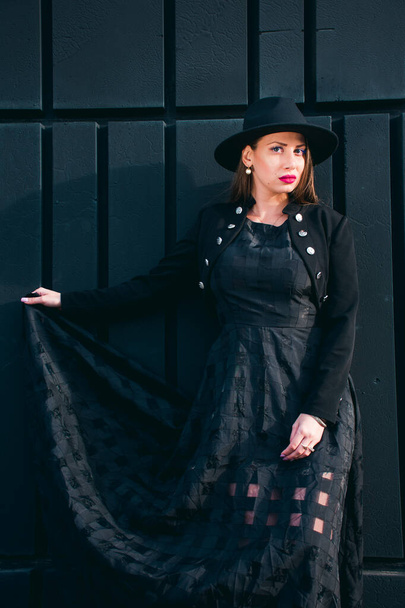 Fotografía de moda con una joven de piel clara con el pelo rubio vestido con un vestido negro largo, torero negro y sombrero posando contra una pared negra. - Foto, imagen