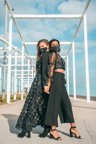 Fashion photography with two young girls posing together, one with fair skin and blonde hair and the other with brown skin with dark hair and both with black protective face masks. - Photo, image