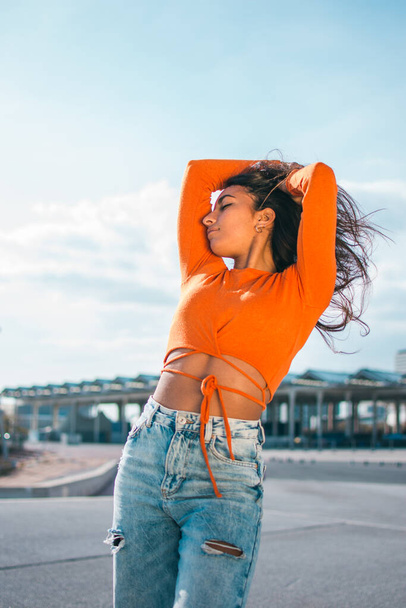 Fashion photography with a slim young woman with dark skin posing. Dressed in an orange top and jeans, on the street during the daylight. - Foto, immagini