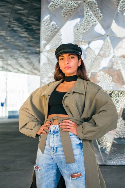 Fashion photography with a young woman with dark skin posing in front of a mirror background. With a black hat and a khaki green jacket. - Foto, immagini