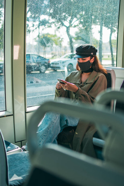 Młoda kobieta o brązowej skórze w czarnej masce ochronnej siedzi w środku transportu publicznego za pomocą telefonu komórkowego.. - Zdjęcie, obraz