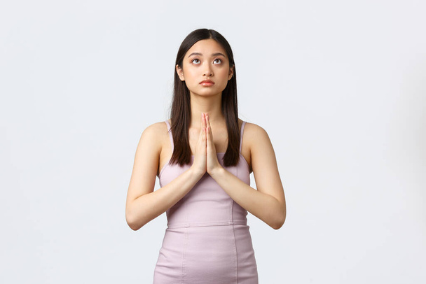 Luxury women, party and holidays concept. Hopeful young asian woman in evening dress, holding hands in plead, begging god, praying and looking up determined, having faith, white background - Photo, image