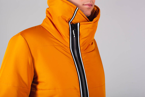 Ein junger Mann europäischen Aussehens in einer orangefarbenen warmen Daunenjacke. Im Rahmen befindet sich ein Kinn mit einer leichten Theke. Die Jacke ist glasiert und der Kragen bedeckt den Hals - Foto, Bild