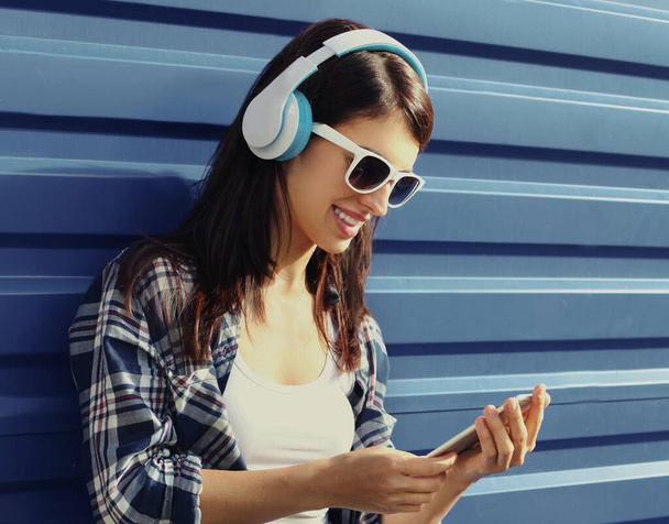 Portrait of young woman in wireless headphones with phone listening to music over a blue background - 写真・画像