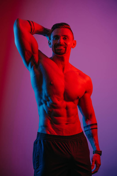 A bodybuilder with a beard is posing with a muscular arm behind his head under the blue and red light. An athletic guy is demonstrating his sporty torso. An athlete in shorts after a workout. - Photo, Image