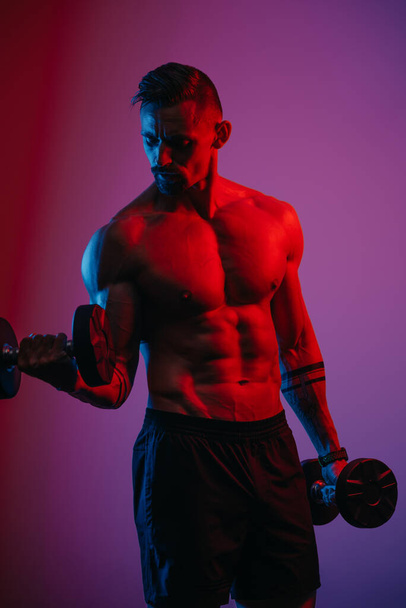 Un homme musclé avec une barbe qui fait des boucles biceps avec des haltères sous les lumières bleues et rouges. Un sportif fait la démonstration de son physique sportif. Un athlète s'entraîne dur les bras. - Photo, image