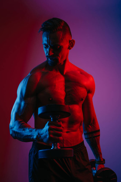 A muscular man with a beard who is doing bicep hammer curls with dumbbells under blue and red lights. An athletic guy is demonstrating his sporty physique. An athlete is training his arms hard. - Фото, зображення