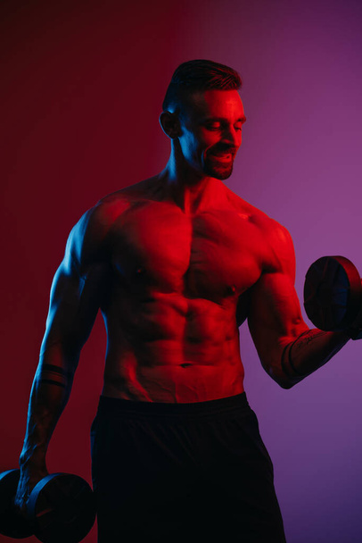 Un hombre musculoso con barba que hace rizos de bíceps con mancuernas bajo luces azules y rojas. Un atlético está demostrando su torso deportivo. Un atleta está entrenando sus brazos.. - Foto, Imagen