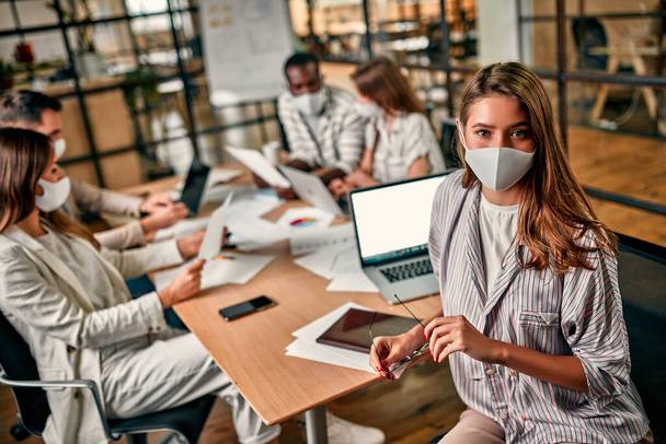 Mulher de negócios caucasiana jovem em uma máscara protetora senta-se em um laptop, segura óculos em sua mão e trabalha com sua equipe ou colegas em um escritório em condições de quarentena. - Foto, Imagem