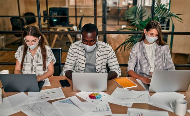 Office work in a pandemic and quarantine. A young group of multi-ethnic business people in protective masks are working on laptops in a modern office. - Photo, Image