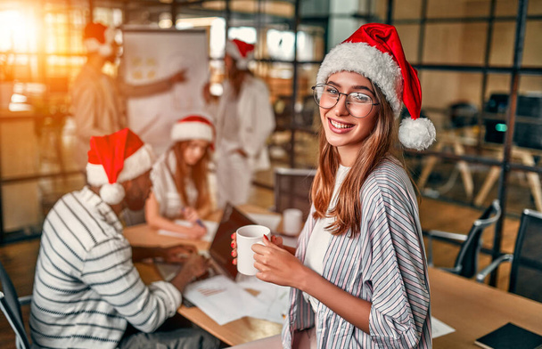 Boldog Új Évet és Boldog Karácsonyt! Multiracionális fiatal kreatív emberek dolgoznak a modern irodában Mikulás sapkában. Jóképű fiatal nő egy csésze kávéval az utolsó munkanapon.. - Fotó, kép