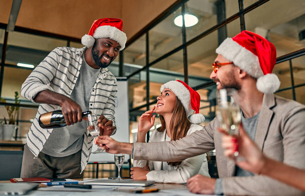 Veselé Vánoce a šťastný nový rok! Multiraciální mladí tvůrčí lidé slaví dovolenou v moderní kanceláři. Skupina mladých podnikatelů pije ve spolupráci šampaňské. - Fotografie, Obrázek