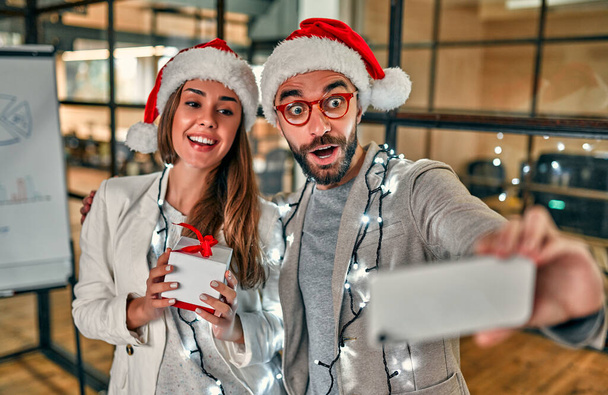 ハッピーニューイヤーとメリークリスマス!サンタの帽子の2人の若い創造的な人々は贈り物を交換し、最後の作業日にスマートフォンを自撮ります. - 写真・画像