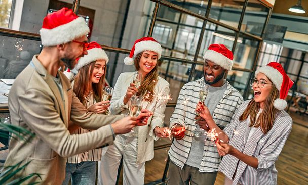 Merry christmas and a happy new year! Multiracial young creative people celebrate the holiday with sparklers in a modern office. A group of young business people are drinking champagne in the coworking space. - Photo, Image