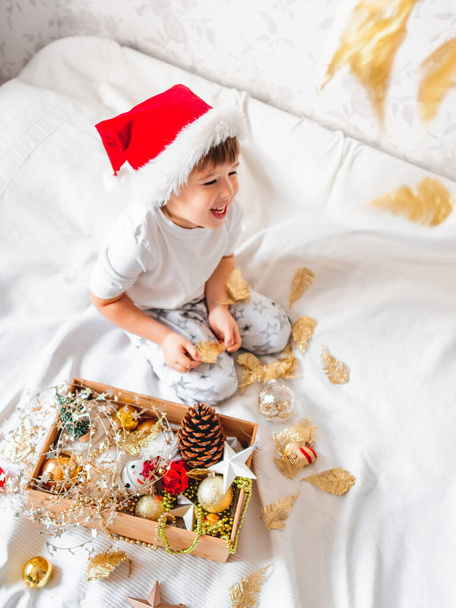 Joyful boy in Santa Claus's hat is playing with decorations for Christmas tree. Funny kid is ready for New Year celebration. Cozy home. Winter holiday spirit. - Photo, Image
