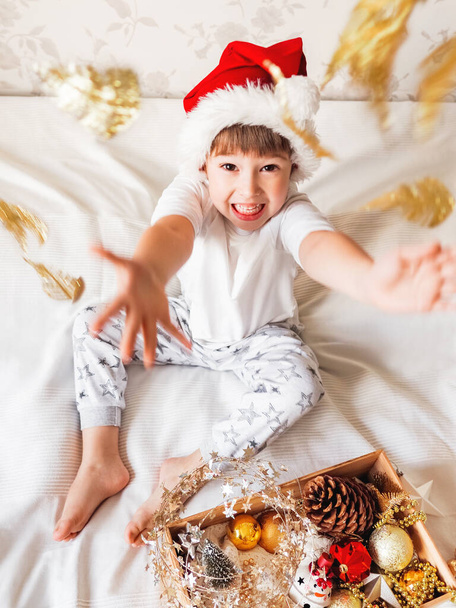 Joyful boy in Santa Claus's hat is playing with decorations for Christmas tree. Funny kid is ready for New Year celebration. Cozy home. Winter holiday spirit. - Foto, imagen