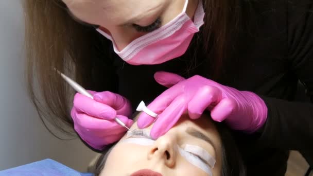 Egy fiatal lány arca egy modern szempilla laminálási eljárás előtt egy professzionális szépségszalonban. A mester speciális ragasztót alkalmaz a szempilla curling eljárás előtt rózsaszín gumikesztyűben és maszkban. - Felvétel, videó