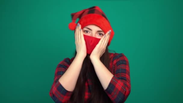 νεαρή γυναίκα σε Σάντα καπέλο και κόκκινη ιατρική μάσκα με σοκαρισμένη έκφραση κρατά τις παλάμες στο μάγουλο - Πλάνα, βίντεο